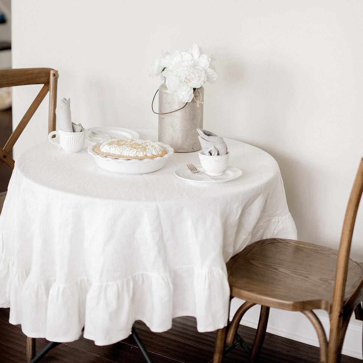 Round table with a white tablecloth, pie, teapot, and flowers in a room.