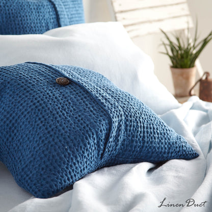 Blue textured pillow on a bed with white bedding and a plant in the background.