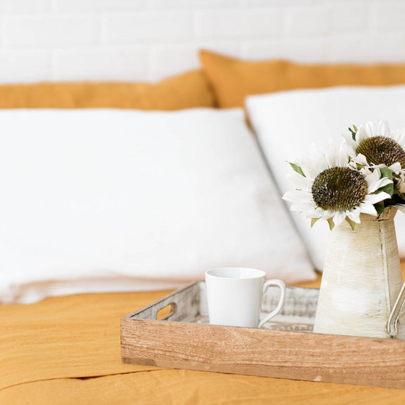 Wooden tray with a mug and vase of flowers on a bed with yellow and white bedding.