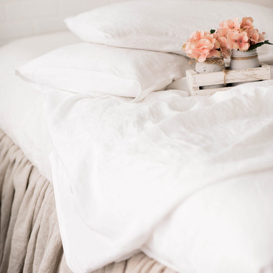 Neatly made bed with white duvet and beige blanket, featuring a small table with flowers on a light background.