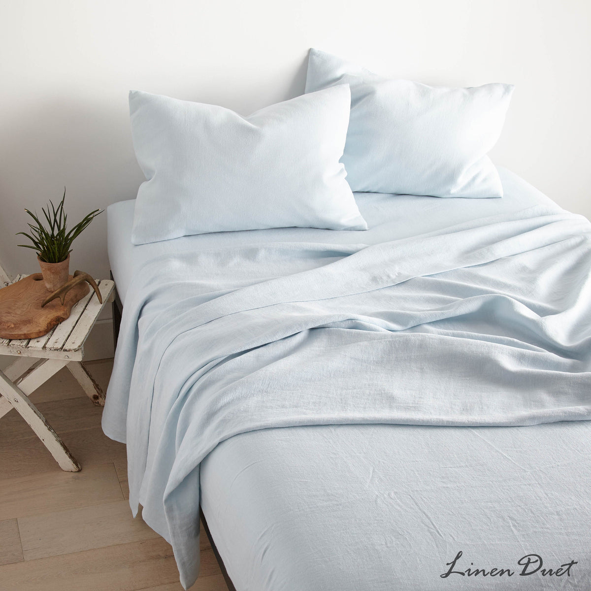 Light blue bedding set on a bed with a small wooden side table and plant in the background.