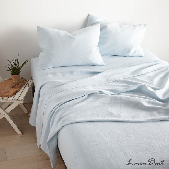 Light blue bedding set on a bed with a small wooden side table and plant in the background.