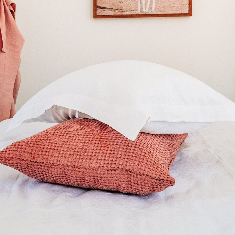 Red textured pillow on a bed with white bedding and a neutral wall.