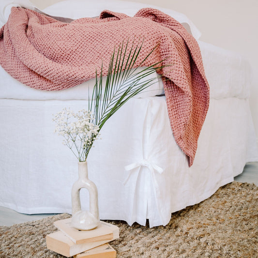 Pink blanket draped over a white bed with a vase of flowers on a wooden block.