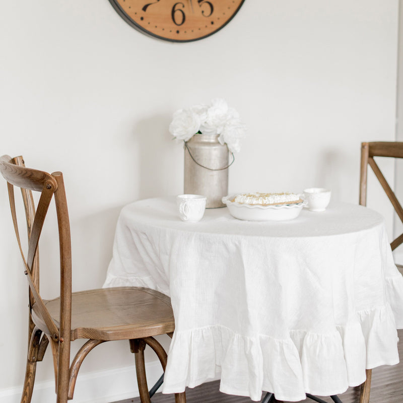 Round table with a white tablecloth, wooden chairs, and decorative items in a room.