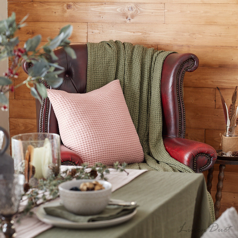 Red leather armchair with pink pillow and green blanket, set against a wooden wall with a table in front.
