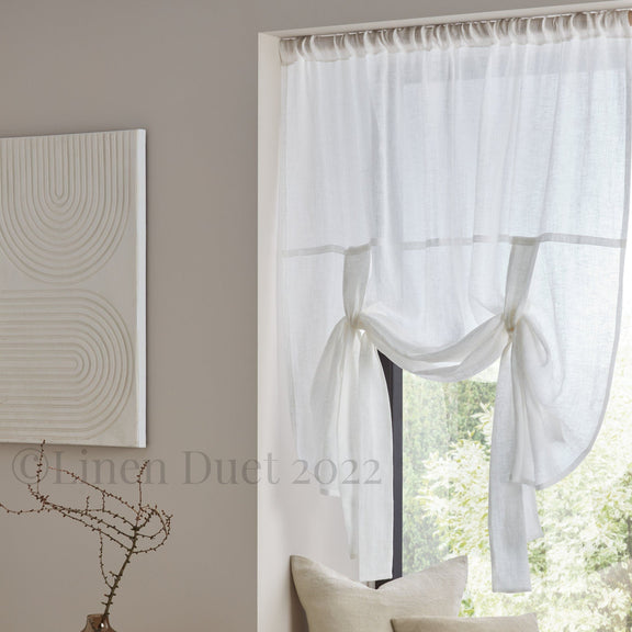 White curtains with a subtle pattern, beige pillows, and a vase on a small table in a room.