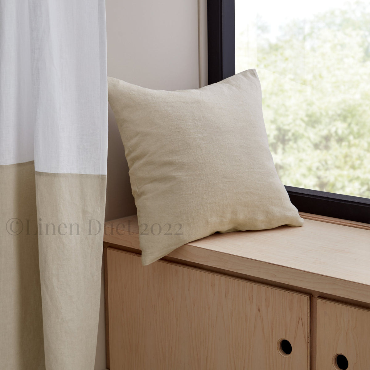 Beige pillow on a wooden window seat with white curtains and a blurred outdoor view.