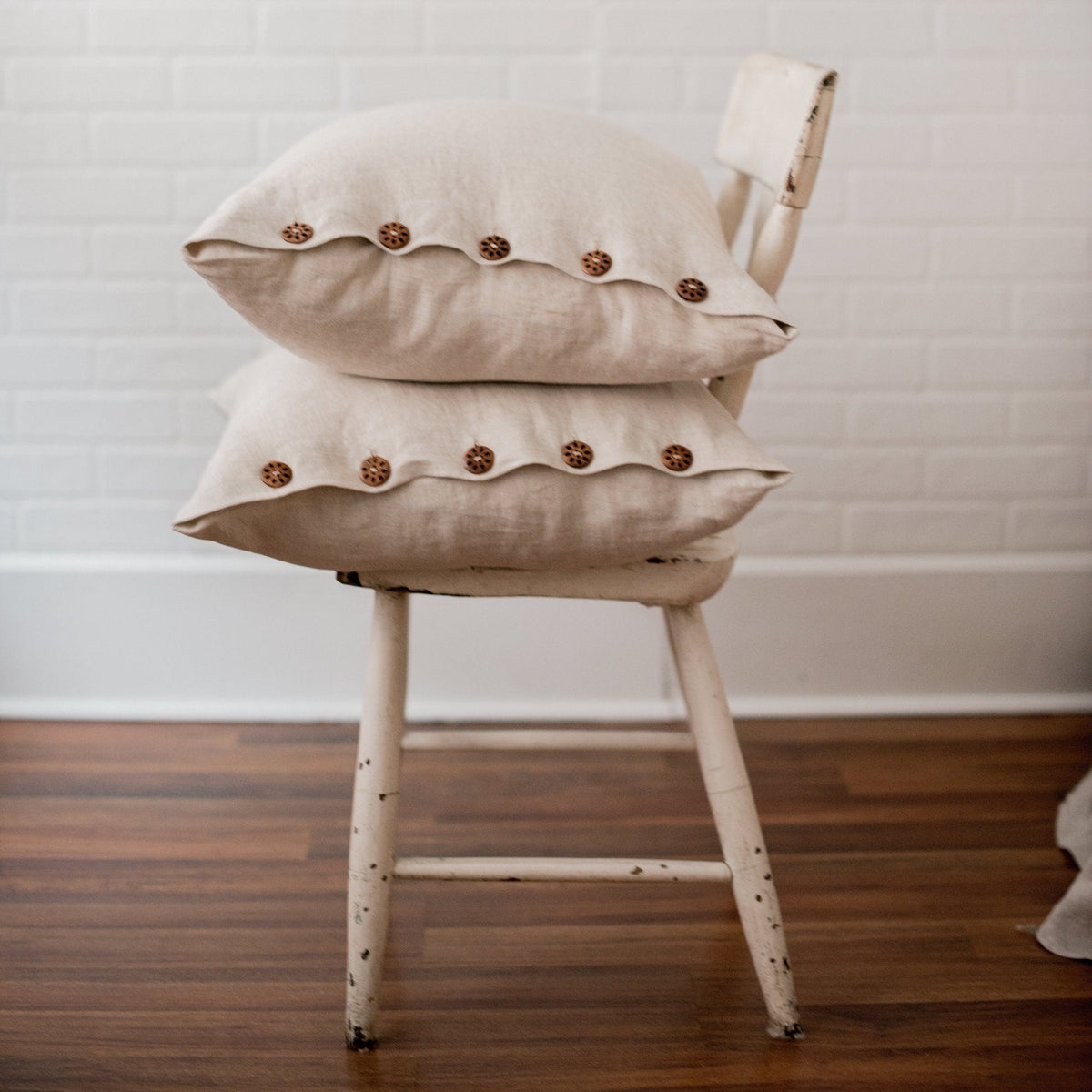 Two beige pillows with button details stacked on a wooden chair against a white brick wall.
