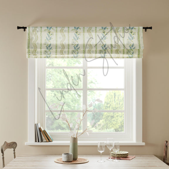 Window with a green and white patterned roman blind, books, a vase, and glasses on a table.